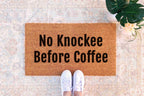 a pair of white sneakers standing on a patterned rug with a rectangular door mat that reads "No Knockee Before Coffee".
